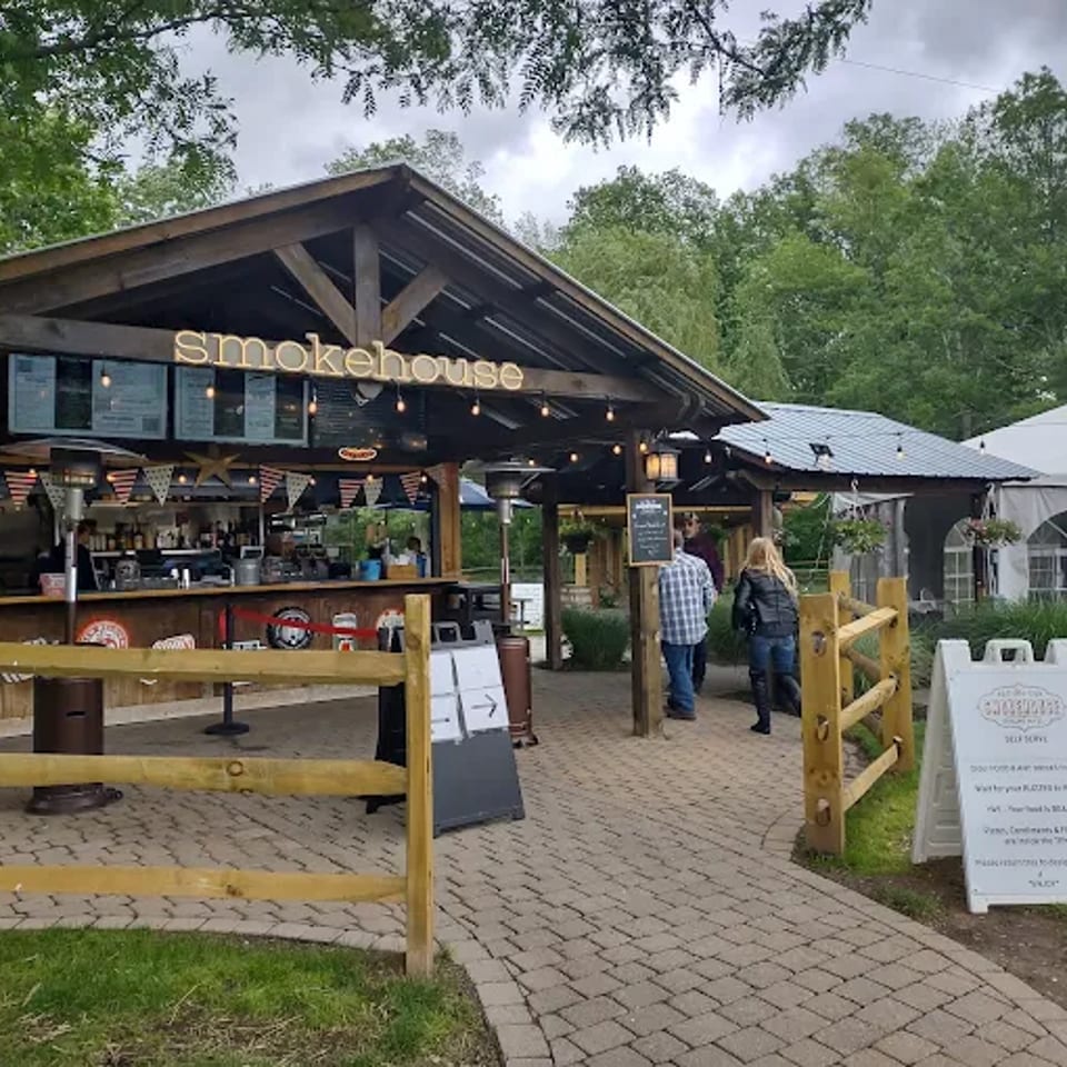 Rustic outdoor smokehouse stand with menu boards, string lights, picnic seating, and visitors beside a white event tent.