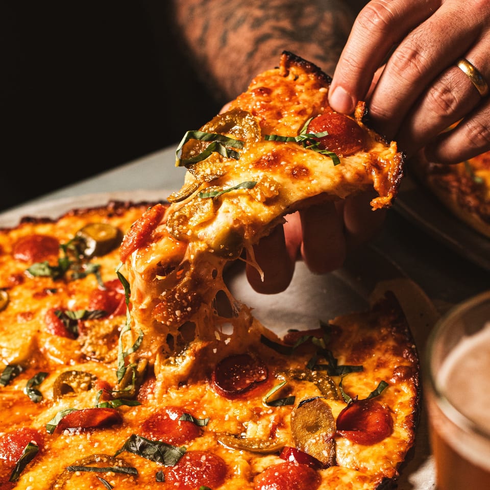 Hand lifts a cheesy pepperoni pizza slice topped with basil and jalapeños from a whole pizza.