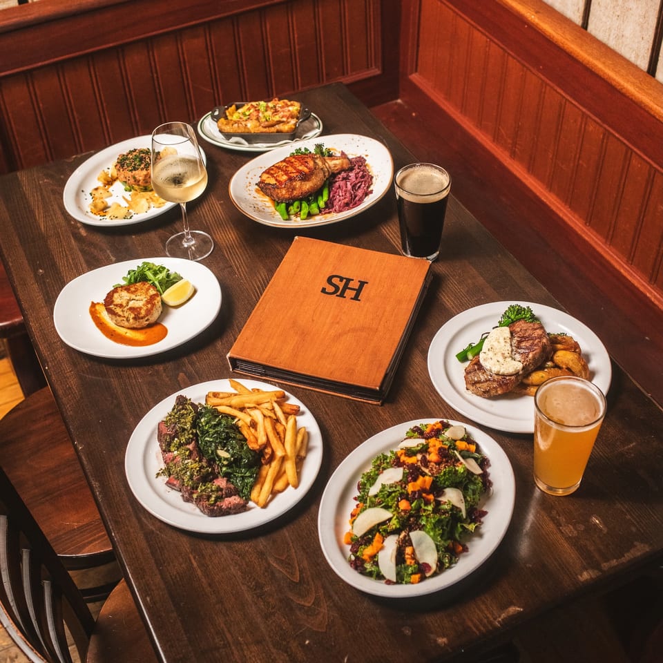Restaurant table filled with plated steaks, roasted vegetables, fries, salads, wine, and beer in a warm wood-paneled booth.