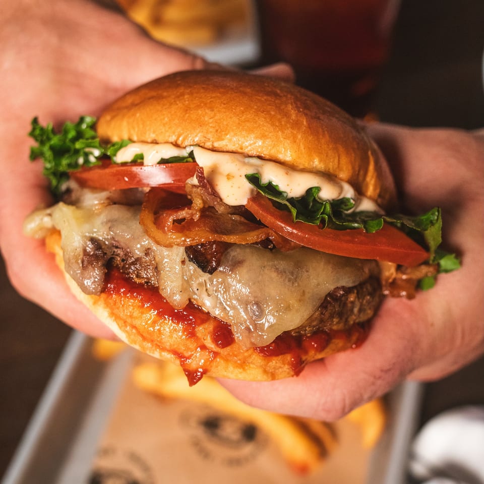 Close-up of a loaded cheeseburger with bacon, lettuce, tomato, crispy onions, and creamy sauce on a wooden table.