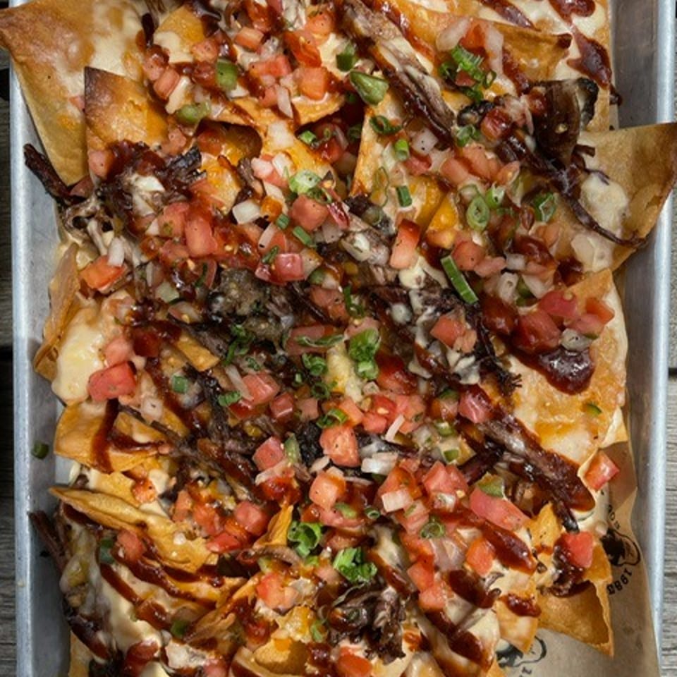 Loaded nachos topped with shredded meat, melted cheese, diced tomatoes, onions, green peppers, and drizzled sauces on a baking tray.
