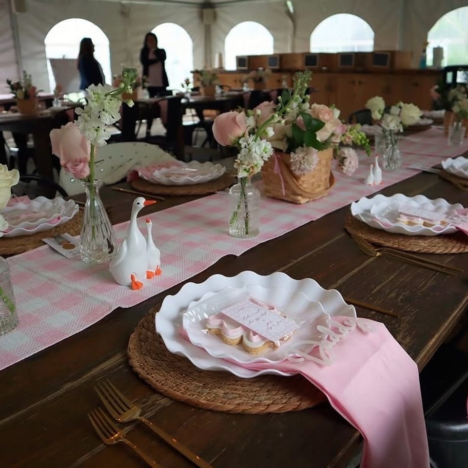 Long dining table decorated with pink runners, floral arrangements, swan figurines, wicker placemats, and elegant place settings inside a tented venue.
