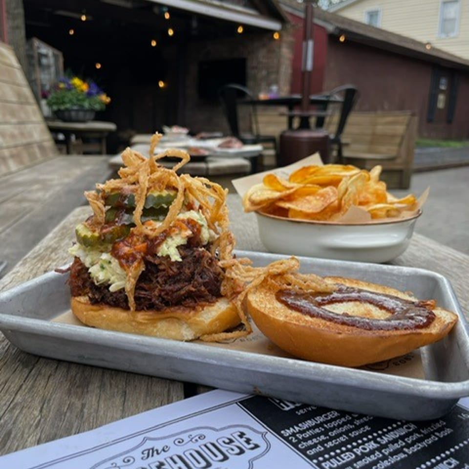 Barbecue sandwich topped with crispy onions beside potato chips on a tray at an outdoor table near a rustic smokehouse.
