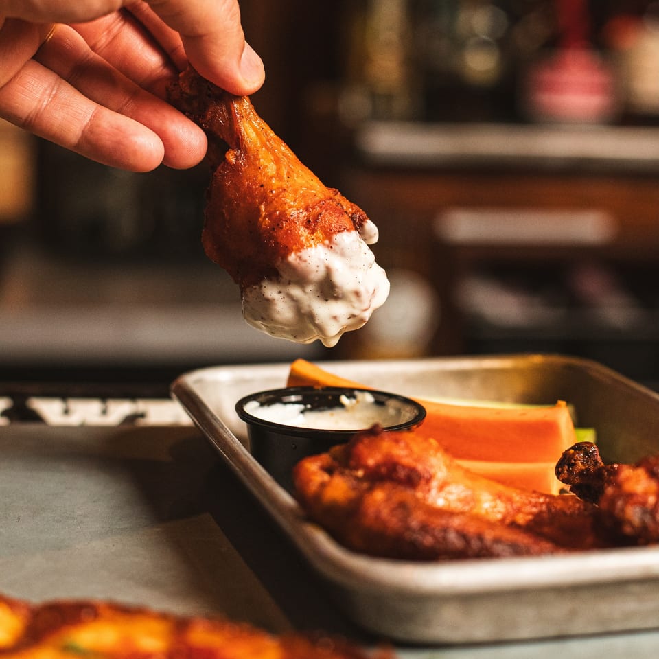 Hand holds a saucy fried chicken wing over a tray of wings and dipping sauce in a warmly lit restaurant kitchen.