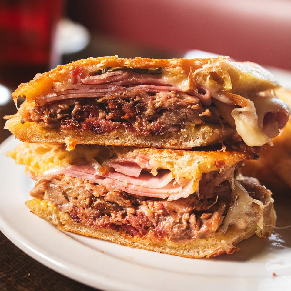 Close-up of a stacked toasted sandwich filled with sliced meats, melted cheese, and crispy layers on a white plate.
