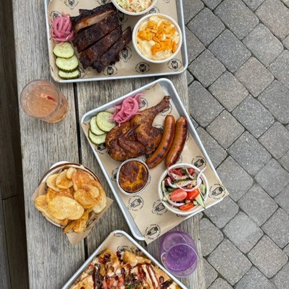 Overhead view of barbecue platters with ribs, chicken, sausages, slaw, pickles, nachos, chips, dipping sauces, and colorful drinks.