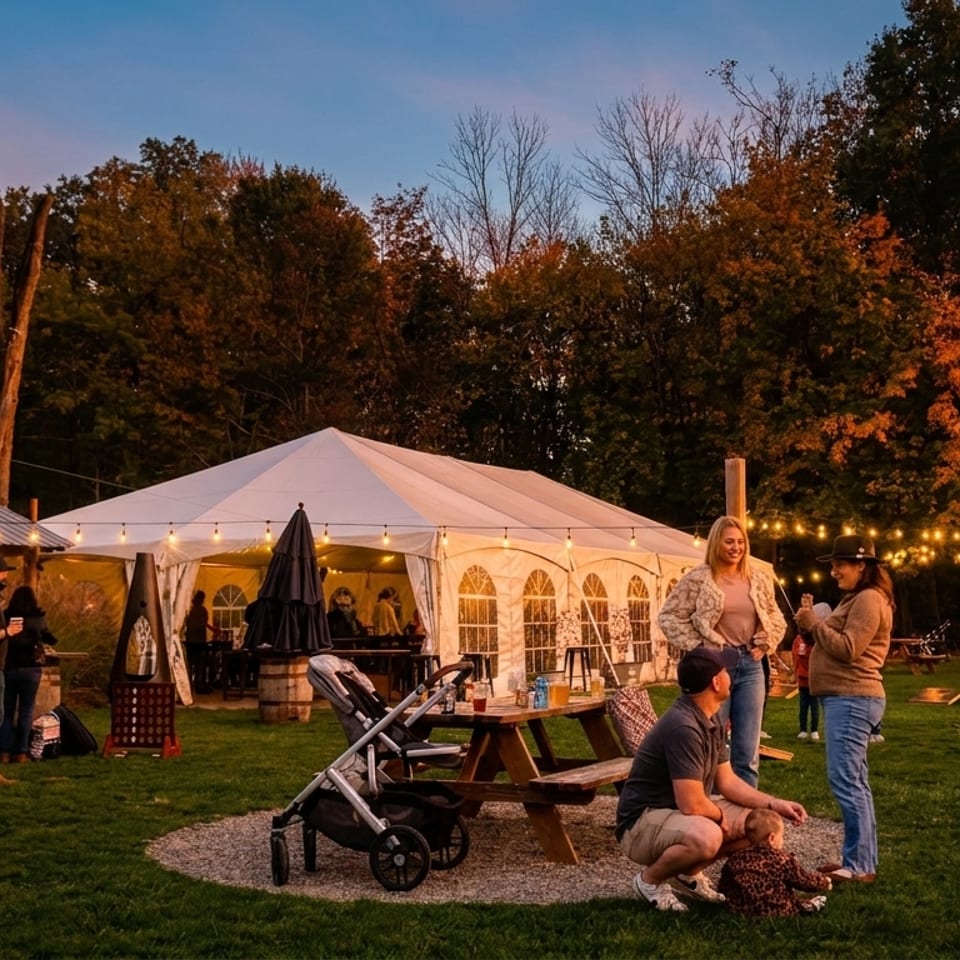 Guests gather on a grassy lawn with string lights, a large tent, and autumn trees at dusk.