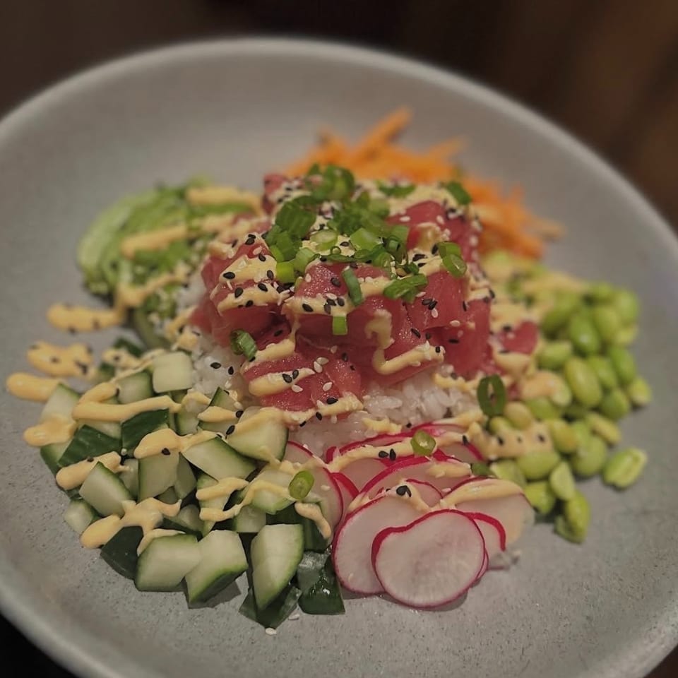 Tuna poke bowl with rice, cucumber, avocado, edamame, radishes, carrots, sesame seeds, scallions, and creamy sauce.