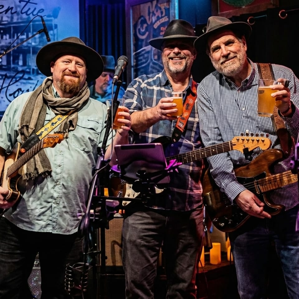 Three men in hats holding beers and guitars, smiling on a dimly lit stage with music equipment.
