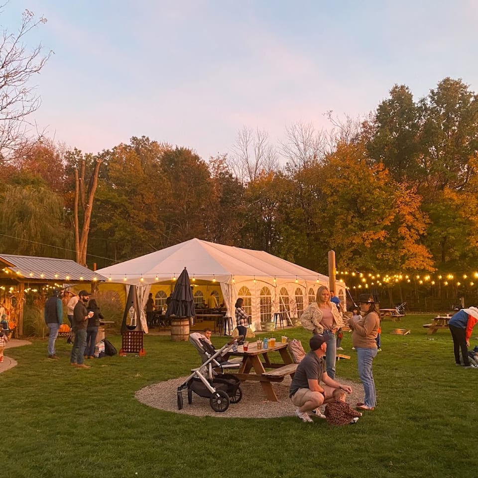 Outdoor restaurant gathering with picnic tables, string lights, a white tent, families, and colorful autumn trees.