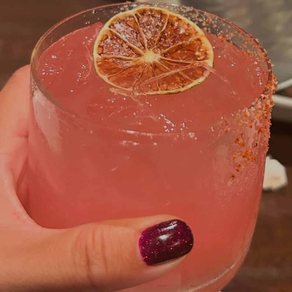 Pink cocktail in a sugar-rimmed glass, garnished with a dried citrus wheel and held by a hand.