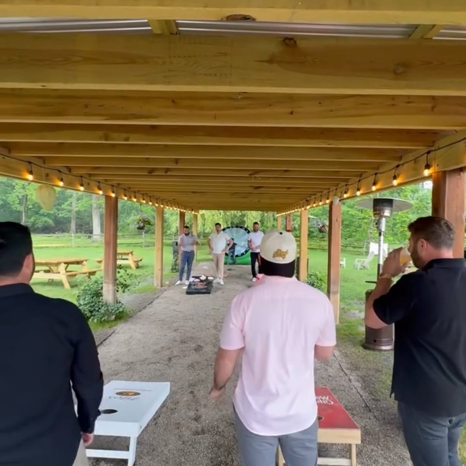 Guests play cornhole beneath a string-lit wooden pavilion overlooking a green outdoor dining area.