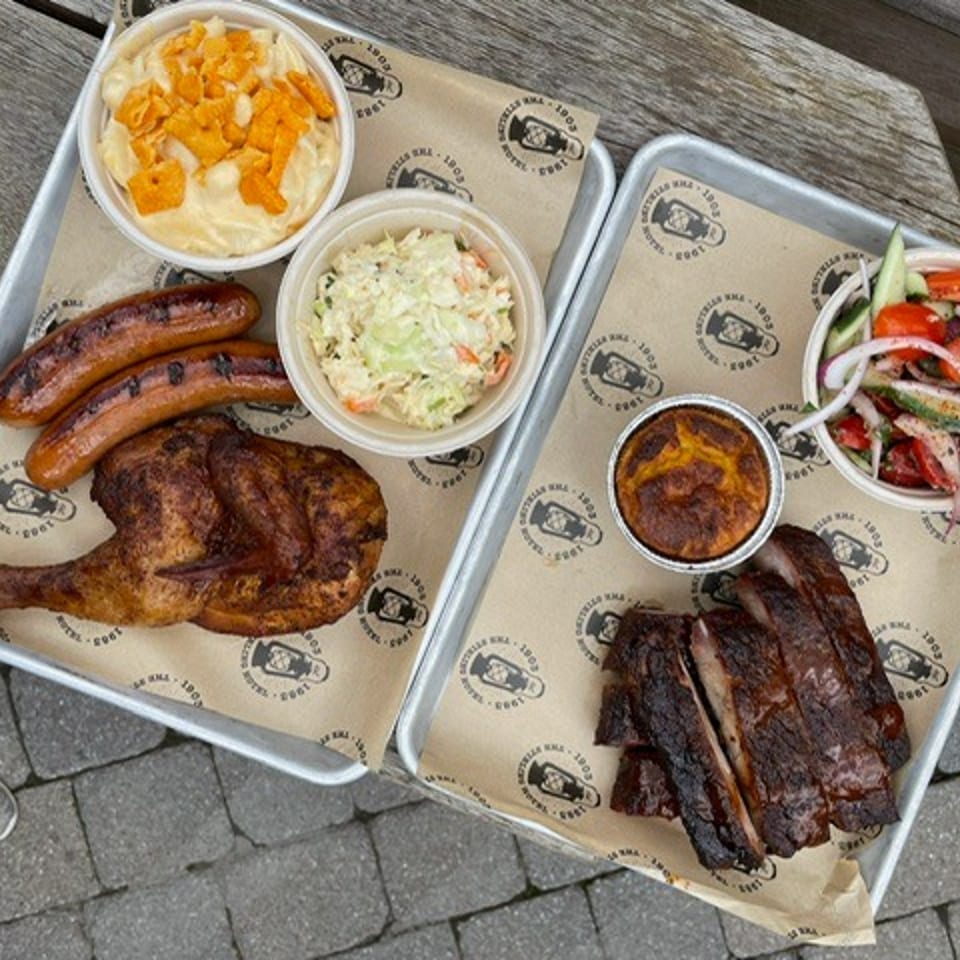 Two trays hold grilled sausages, roast chicken, ribs, coleslaw, macaroni salad, cornbread, and a fresh tomato-cucumber salad.