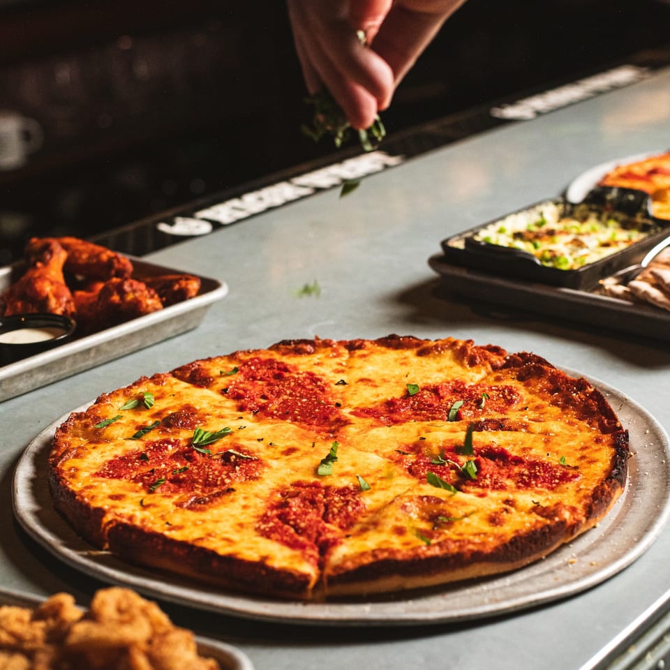 Cheesy rectangular pizza garnished with basil sits on a serving tray amid assorted dishes while a hand adds herbs.