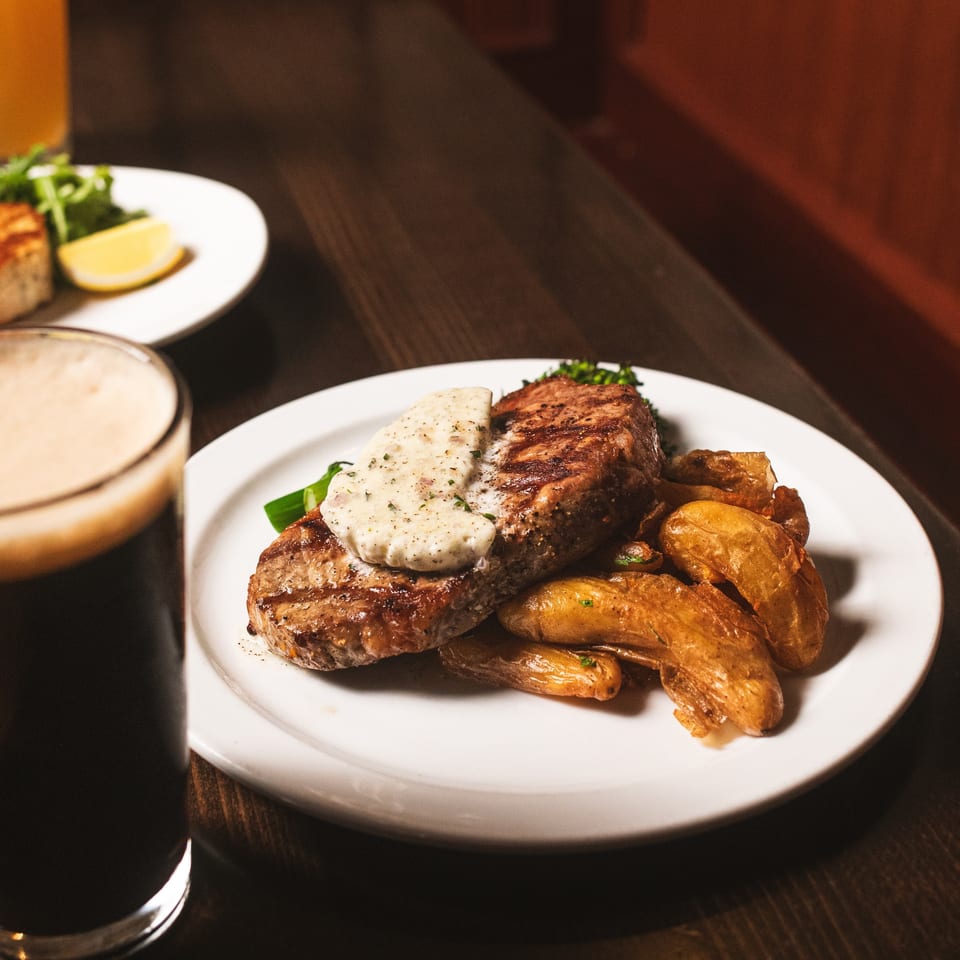 Grilled steak topped with creamy herb sauce, roasted potatoes, and vegetables on a white plate at a warmly lit restaurant table.