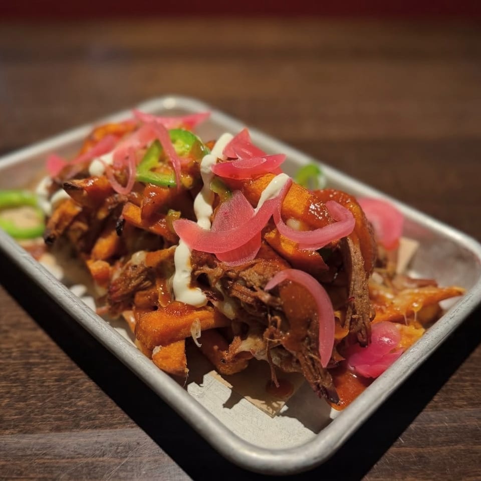 Loaded fries topped with shredded meat, barbecue sauce, white crema, pickled onions, and jalapeños in a metal tray.